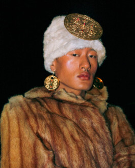 Model Sachit Gurung wears a vintage fur coat, ornate golden earrings, and a white faux fur hat adorned with a large gold medallion, set against a stark black backdrop. 