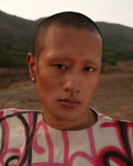 Close-up outdoor portrait of model Sachit Gurung wearing a graffiti-style shirt with pink and black patterns, standing against a muted mountain landscape during golden hour. 
