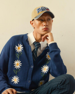 Sachit Gurung seated in a relaxed pose wearing a daisy-embroidered blue cardigan, a striped tie, collared shirt, and beige baseball cap against a plain backdrop. 