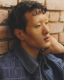 Close-up of model Sachit Gurung leaning pensively against a rustic brick wall, wearing a textured navy jacket with fine white pinstripes. 