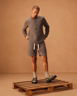 Model Sachit Gurung poses on a wooden pallet in a minimal beige-toned studio, wearing a grey co-ord set with a Henley-style top, matching shorts, grey socks, and sandals. 