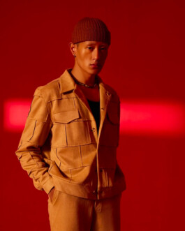 Model Sachit Gurung stands against a vivid red backdrop wearing a structured camel-toned jacket and matching trousers, accessorized with a beanie and necklace. 