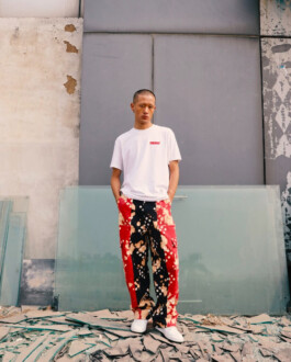 Sachit Gurung stands on broken glass and rubble in front of a raw industrial wall, wearing a white t-shirt and tie-dye red, black, and beige trousers. 