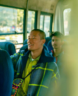 Model Sachit Gurung smiling while sitting on a bus, wearing a vibrant checkered Péro overcoat layered over a yellow striped shirt, with a colorful woven crossbody bag. 