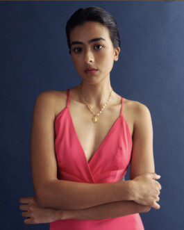 Model Medhavi Chettri in a pink strappy dress paired with a gold sun pendant necklace, styled for Olio campaign against a navy blue backdrop 