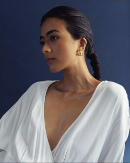 Model Medhavi Chettri wearing a white draped blouse and chunky gold hoop earrings against a deep blue background for Olio campaign 