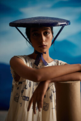 Akriti Jha wearing a cream dress with navy blue folk-inspired illustrations and a wide sculptural hat, shot in front of a dreamy blue-toned backdrop for Ahmev campaign 