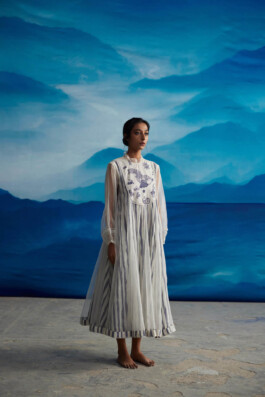 Akriti Jha wearing a sheer white organza dress with hand-drawn indigo motifs and vertical stripes, barefoot against a blue mountain-inspired backdrop for Ahmev SS24 campaign 