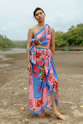 Merrylin Boro stands barefoot on a beach wearing a vibrant pink and blue Verandah outfit with floral and leopard motifs 