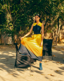 Model Akriti Jha posing in a yellow and blue gradient dress for Bodice, standing between two chairs in natural sunlight 