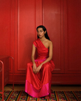 Akriti Jha in a red Raw Mango silk saree-inspired ensemble, seated against a red paneled wall 