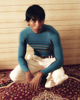 Gaurav Gharu in a fitted blue striped top and cream trousers, crouched on a patterned carpet in front of wood-paneled walls 