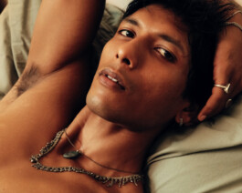 Close-up of Gaurav Gharu lying down shirtless, wearing silver rings and layered necklaces with a calm, intense gaze 