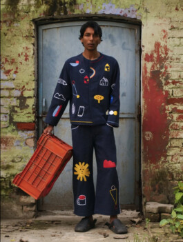 Model Gaurav Gharu wearing a navy blue co-ord set with colorful embroidered motifs, holding an orange crate for Fenmy Taxena fashion campaign 