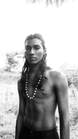 Black and white portrait of male model Savio Fedrick standing shirtless outdoors, wearing statement pearl jewelry 