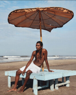 Savio Fedrick sitting shirtless on a blue wooden bench under a beach umbrella with waves in the background 