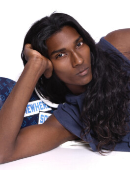 Savio Fedrick with long hair lying on a white surface in a studio wearing a blue outfit 