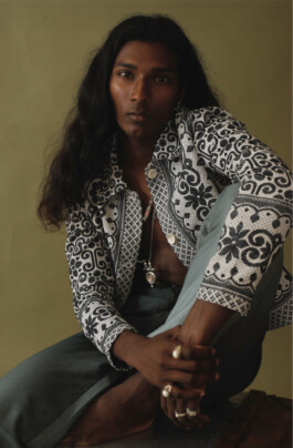 Savio Fedrick, ALF model, sits barefoot in a patterned black and white jacket and green trousers, styled with rings and layered necklaces 