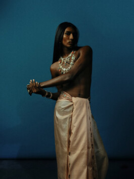 Model Savio Fedrick from ALF modeling agency wearing traditional draped attire and statement jewelry in an artistic editorial photoshoot against a blue backdrop 