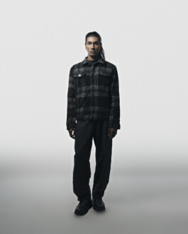 Full-length editorial photo of model Savio Fedrick standing against a minimal light-grey backdrop wearing an all-black outfit and plaid jacket. 