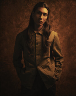 Moody editorial portrait of model Savio Fedrick standing against a warm textured backdrop wearing an olive jacket with hands in pockets. 