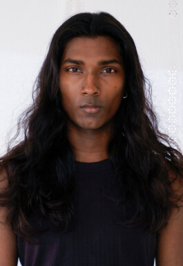 Savio Fedrick stares directly into the camera with long wavy hair worn loose, dressed in a simple black ribbed tank against a soft white backdrop. 