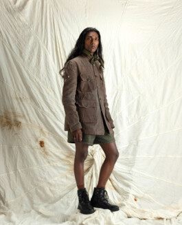 Model Savio Fedrick standing against a textured white backdrop, wearing a rugged brown utility jacket, olive green textured shorts, and black combat boots, styled with a tied olive neck scarf. 
