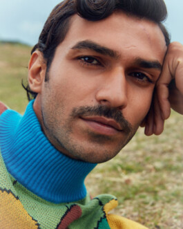 Close-up portrait of a male model with styled hair and mustache wearing a vibrant blue turtleneck and colorful knitwear, photographed outdoors by Dhruvin Shah 