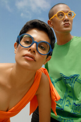 Close-up of two models wearing colorful round glasses under a blue sky, with Sachit Gurung in a green shirt and yellow glasses for Stones 3D campaign 