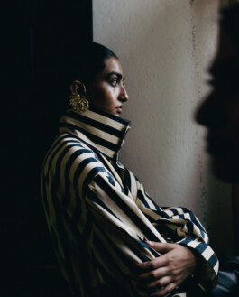 Female model in a striped high-neck outfit with statement gold earrings, seen in profile backstage at Lovebirds 2026 presentation, with soft dramatic lighting. 