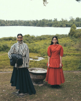 Two female models standing outdoors near a lake, one in a striped oversized top and black skirt, the other in a structured red dress, backstage at Lovebirds 2026 presentation. Represented by ALF modeling agency India. 