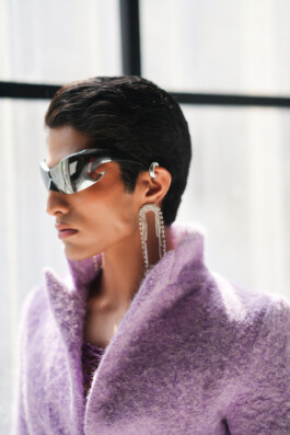Male model Siddharth Raghavan wearing futuristic sunglasses, statement earrings, and a textured lavender coat 