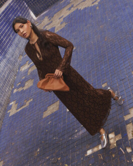 Merrylin Boro wearing a brown lace gown and silver flats, holding a tan Mistry handbag, standing on a tiled blue floor 