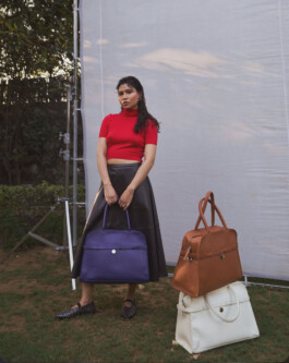 Model Merrylin Boro in red top and black skirt holding a purple Mistry handbag, standing beside stacked brown and white handbags on grass 
