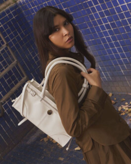Model Merrylin Boro in a brown outfit carrying a white structured Mistry bag in front of a blue-tiled wall 