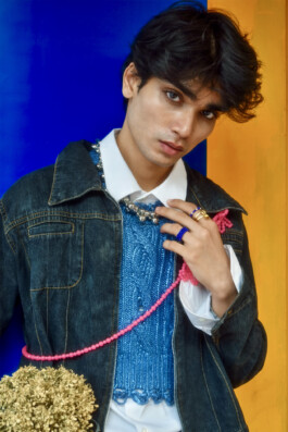 Model Prince wearing blue knit vest, denim jacket, and statement jewelry against a vibrant backdrop 