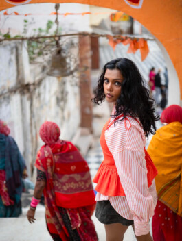 Ishra Philipose in striped shirt and orange top turning back under an arch with women in traditional sarees – Wasala campaign 