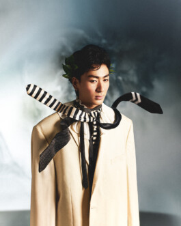 Nam Dorjee wearing a cream blazer and dramatic striped scarves, with laurel leaves in hair, set against an ethereal background 