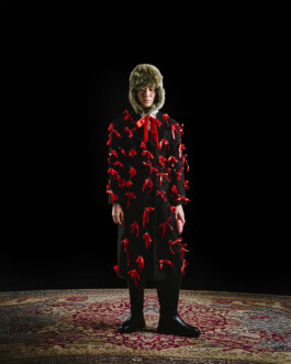 Nam Dorjee in a long black coat adorned with red satin bows and fur hat, standing against a dramatic black background on a patterned carpet 