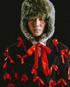 Nam Dorjee wearing a statement coat adorned with red ribbons and a furry winter hat in a high-fashion editorial portrait 