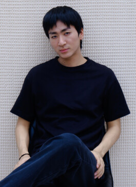Nam Dorjee seated in black t-shirt and navy pants, posing for clean model digitals against a textured white wall 