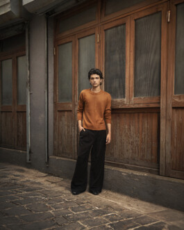 Model Rishabh Dubey standing outdoors in an orange sweater and black trousers during an editorial photoshoot. 