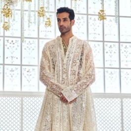 Vikas Mandaliya wearing an ivory lace sherwani by Abu Jani Sandeep Khosla, posing in a regal Indian setting with gold chandeliers and floral décor 