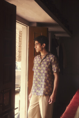Model Siddharth Raghavan stands by an open door, wearing a short-sleeved purple and gold printed shirt, bathed in soft morning light 