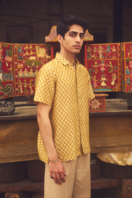 Model Siddharth Raghavan wearing a yellow zigzag patterned shirt standing in front of a traditional red hand-painted wooden panel featuring mythological figures 