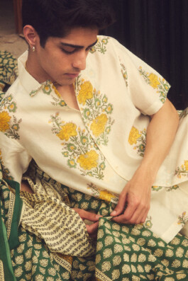 Close-up of model Siddharth Raghavan wearing a white shirt with yellow floral hand-block print, seated on traditional green and cream Indian textiles 