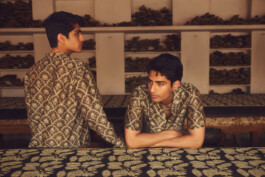 Two male models in hand-block-printed golden shirts posing inside an artisanal Indian textile studio with wooden printing blocks on shelves 