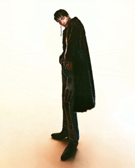 Editorial fashion photo of model Nam wearing studded pants, black boots, and a long fur coat, styled with bold accessories against a minimal background 