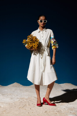 Model Akriti Jha wearing a white button-down dress with sequin-embroidered sleeves, styled with red heels and sunglasses, holding a bouquet of yellow flowers for Mohammed Mazhar campaign 