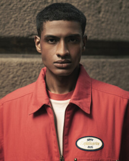 Editorial portrait of model Albhei Gazi wearing a red jacket over a white T-shirt, standing against a textured stone wall. 